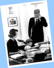 Evelyn Lincoln, President Kennedy's Personal Secretary, looks on while the President takes an important phone call and his son, John F. Kennedy, Jr., plays with her typewriter.